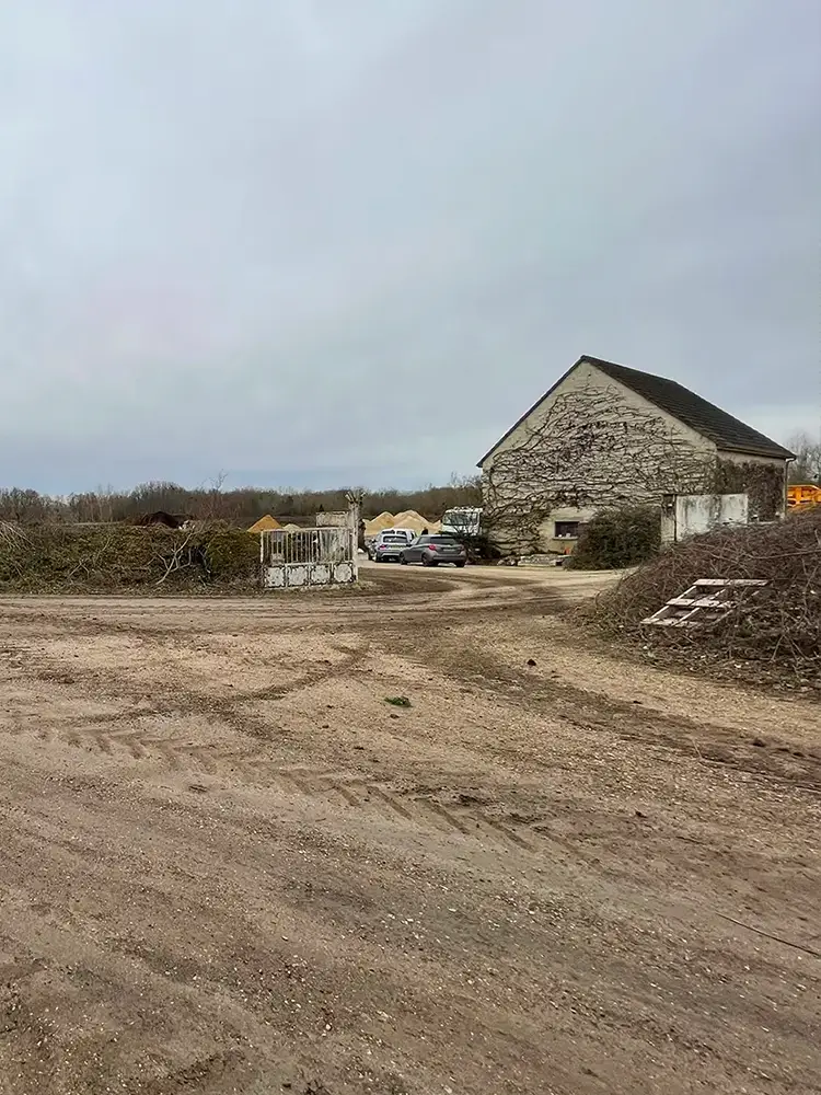 Vue d’une cour gravillonnée avec bâtiment agricole en arrière-plan et plusieurs véhicules stationnés