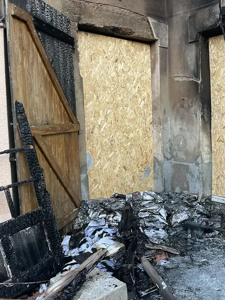 Façade d’un bâtiment calciné avec volets brûlés, fenêtres condamnées et amas de débris carbonisés au sol