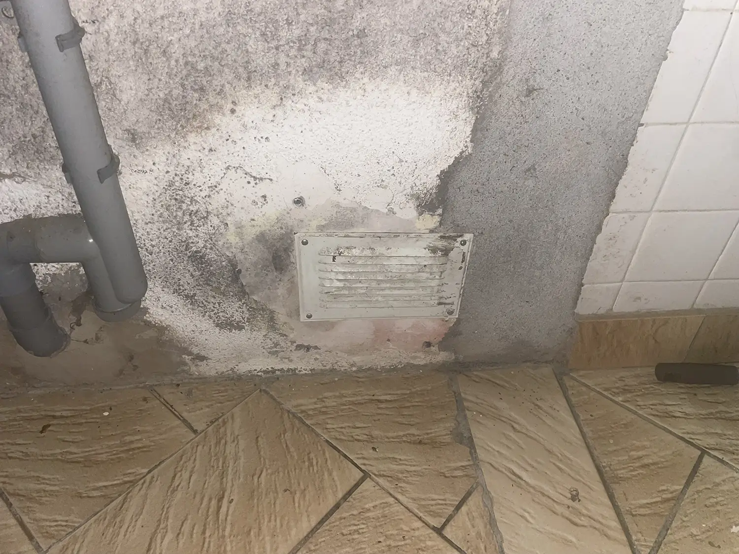 Grille d’aération murale encrassée sur un mur humide avec moisissures et canalisations PVC